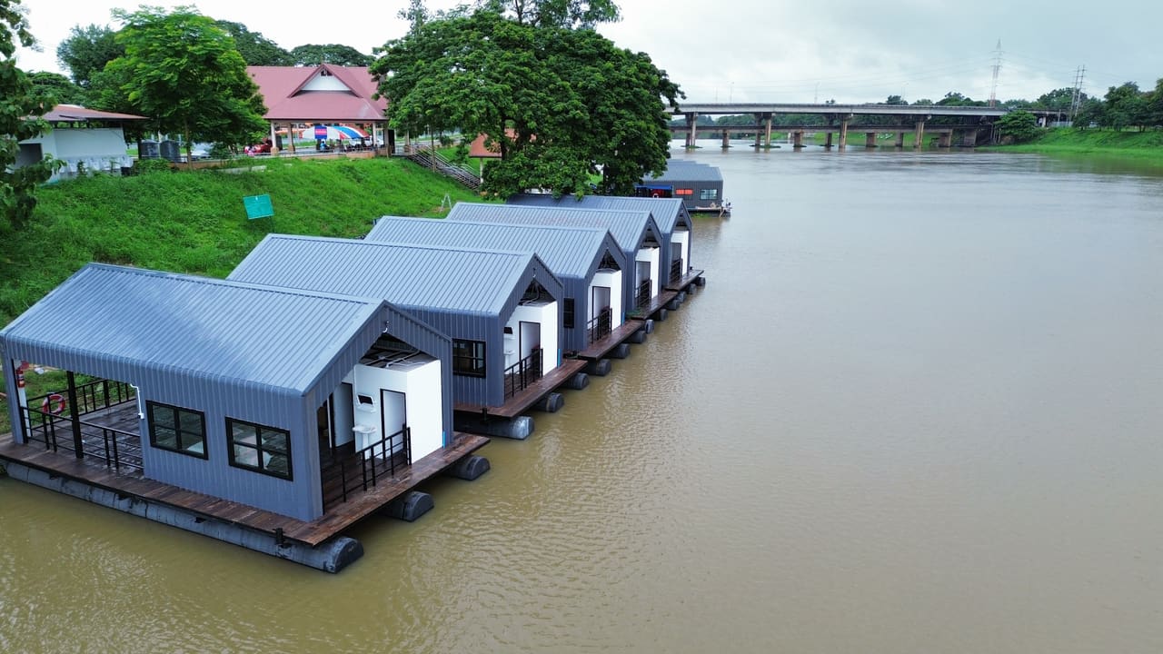 แถวแพที่พักลอยน้ำหลังคาจั่วริมฝั่ง มีสะพานอยู่ด้านหลัง — แพที่พักล่องในลำน้ำ เขื่อน แม่น้ำ T.K.M. Aquatic